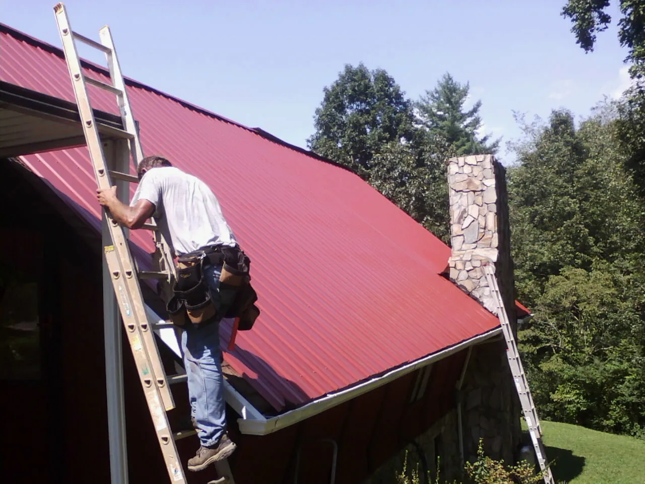 Roofer on a ladder beside a red metal roof and stone chimney — Soffit & Fascia Repair by Roofing Brothers in Ray